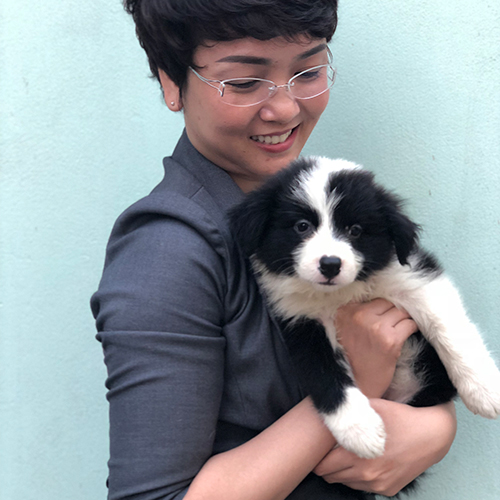 Người phụ nữ mỉm cười ôm chú chó con lông đen trắng, đứng trước nền tường màu xanh.